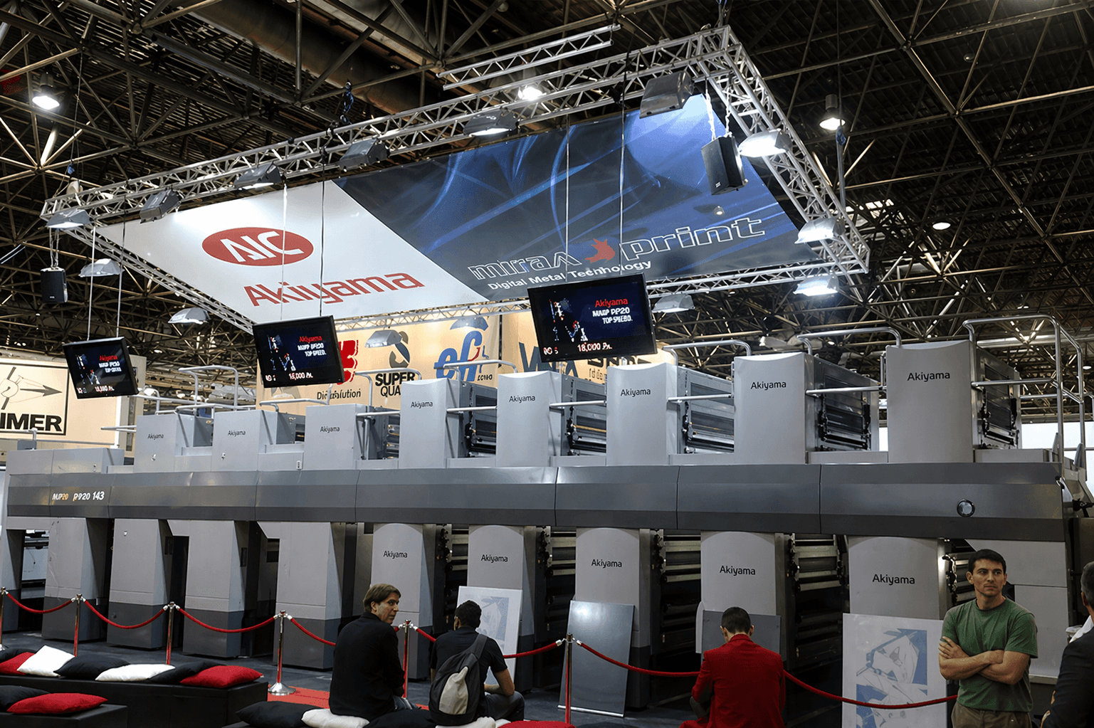 Groot hangend beursbanner in trussconstructie met verlichting boven een Akiyama drukpers op een vakbeurs.