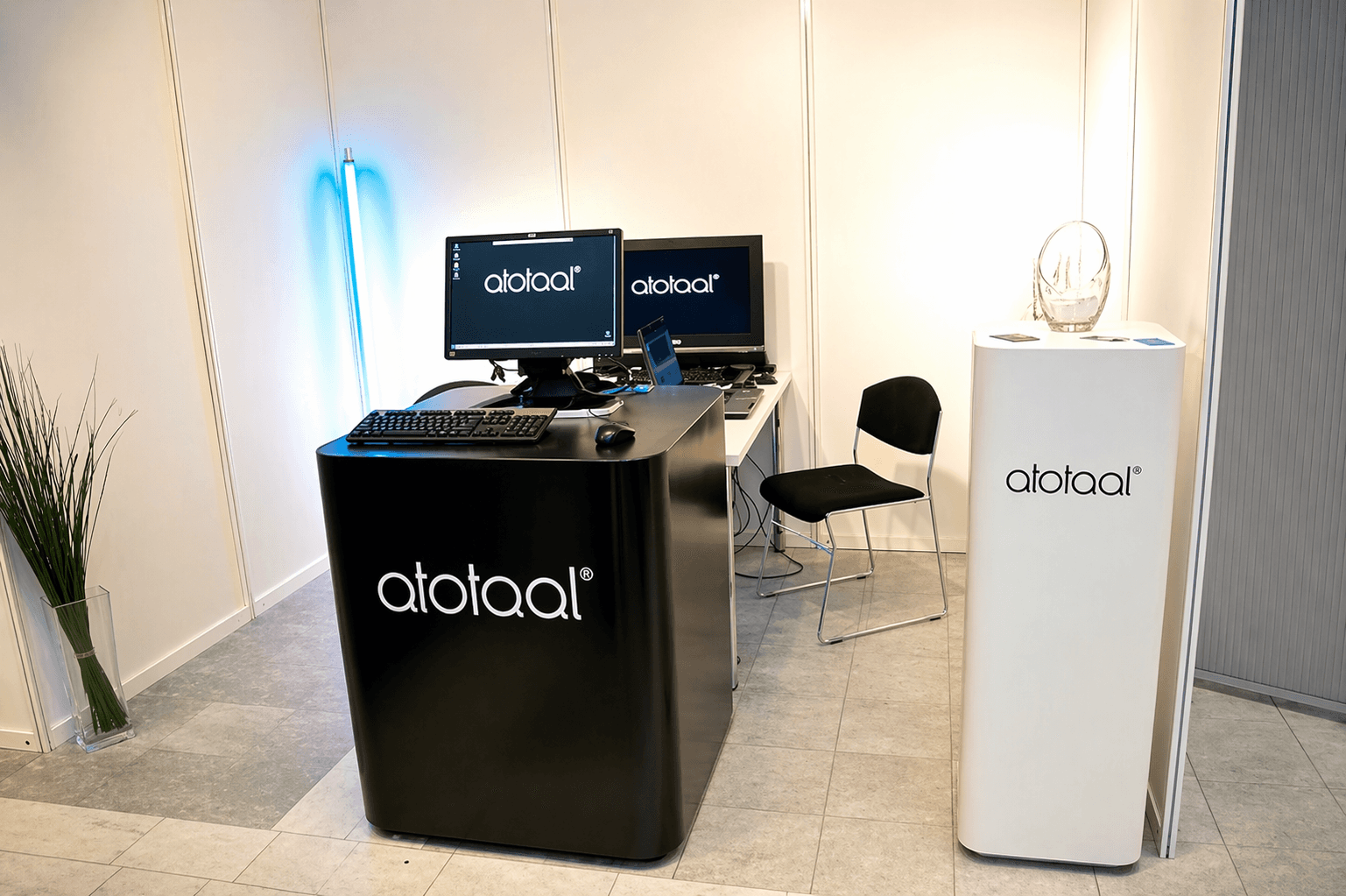 Minimalistische presentatiehoek met zwarte balie en witte displayzuil met atotaal-belettering, computerschermen met logo en strakke kantoorinrichting.