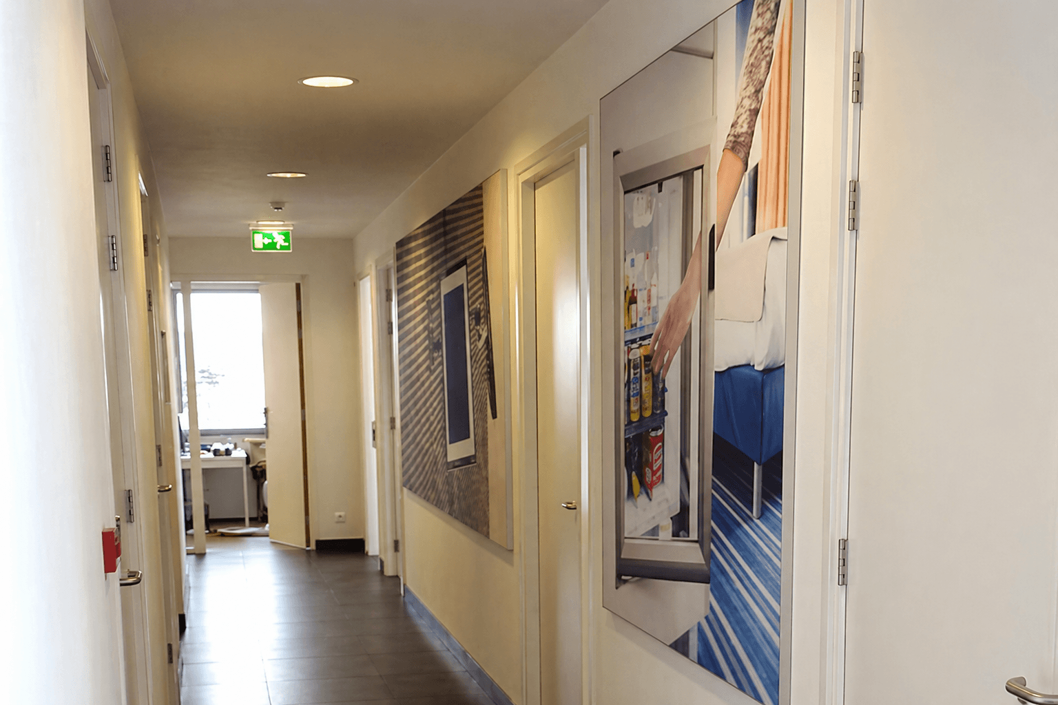 Gang met grote fotoprint-wandpanelen op binnenmuren en verlichte nooduitgangsigning, toegepast als decoratieve interieurbelettering en wayfinding.