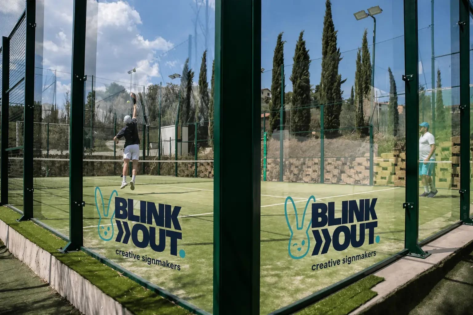 Glaswand van een padelbaan met Blink Out creative signmakers-logo in turquoise en donkerblauw, aangebracht als transparante raamsticker op sportbeglazing.
