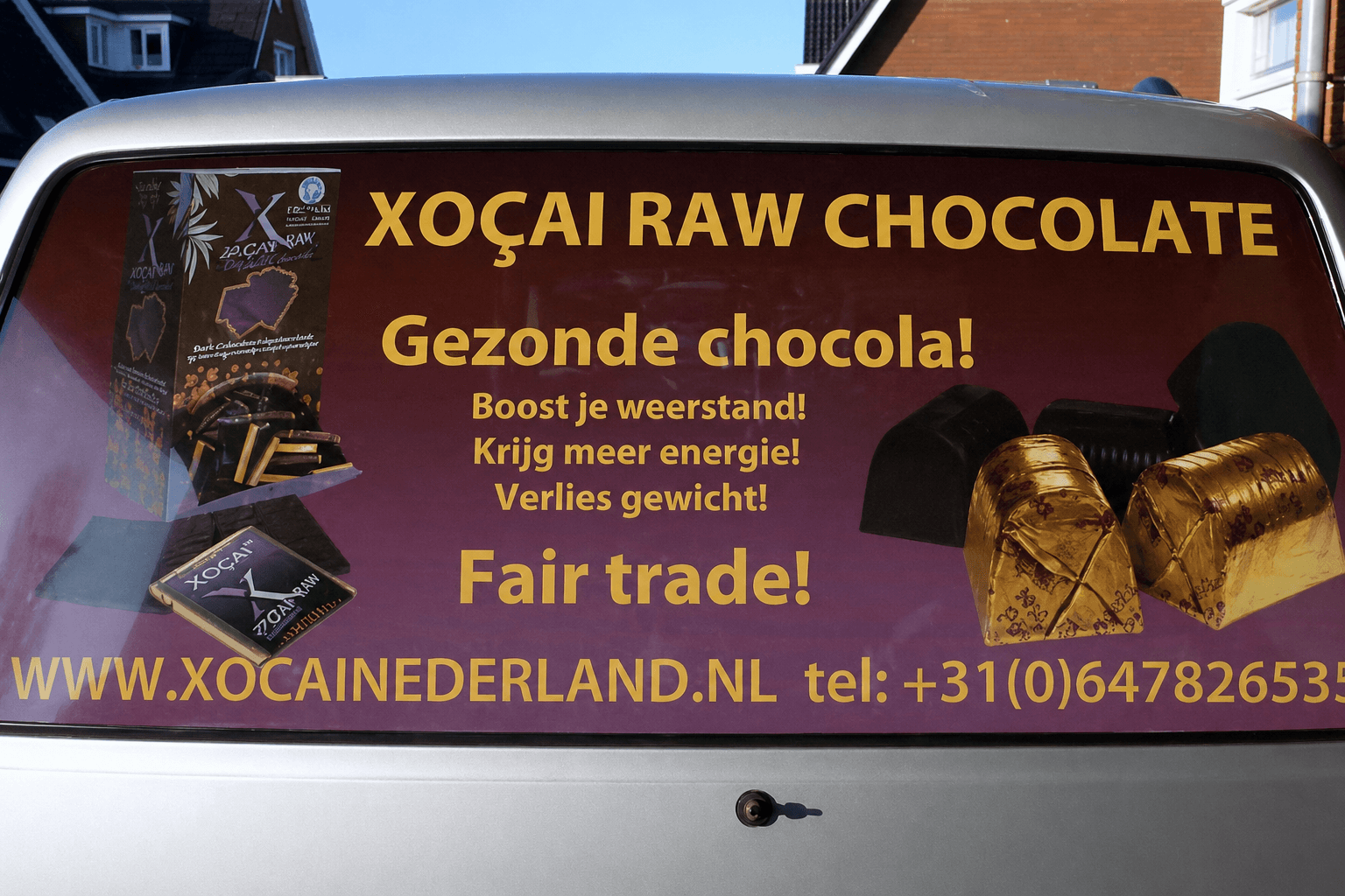 Voertuigbelettering op de achterruit van een zilveren bestelauto met paarse Xoçai Raw Chocolate reclame, gele tekst en chocoladeafbeeldingen.