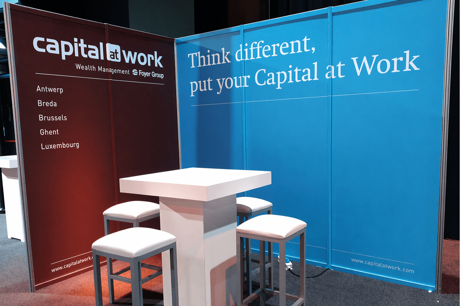Beursstand met rode en blauwe wandpanelen van Capital at Work, grote slogan in witte letters en strakke zitopstelling met hoge tafel en krukken.