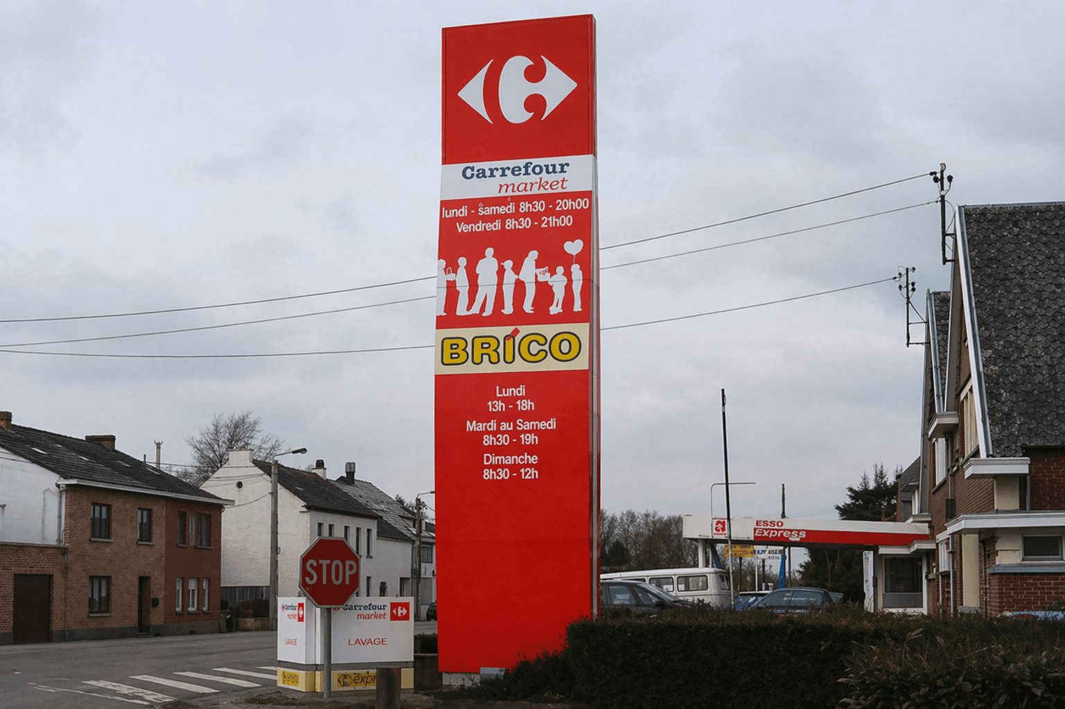 Hoge rode reclamezuil met Carrefour market en BRICO langs een straat bij een winkel- en tankstationlocatie.