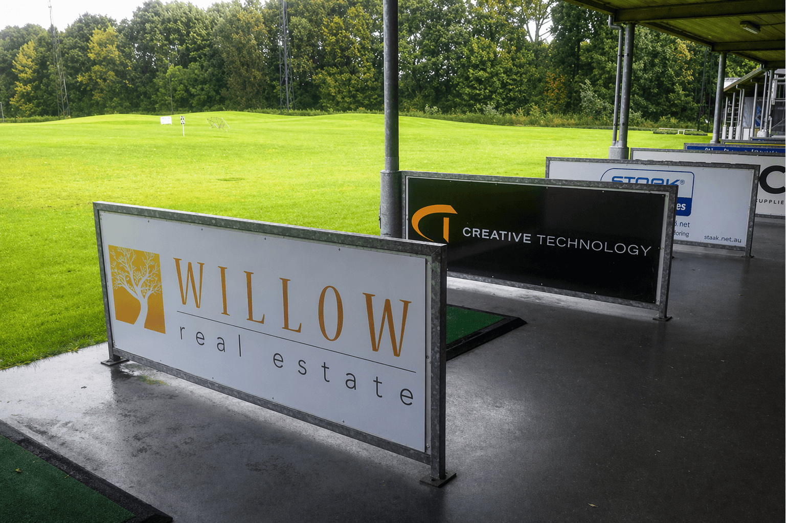 Reclameborden in metalen frames met Willow real estate en Creative Technology langs een overdekte golfafslagplaats bij een groen sportveld.