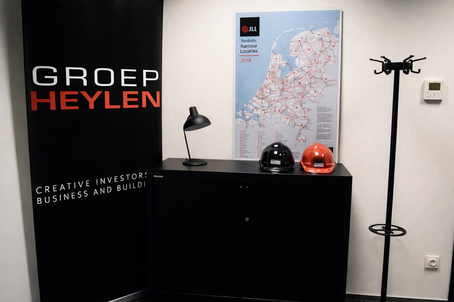 Kantoorinterieur met zwarte roll-up banner van Groep Heylen, wandposter met kaart en branding, en zwarte kast met twee veiligheidshelmen.