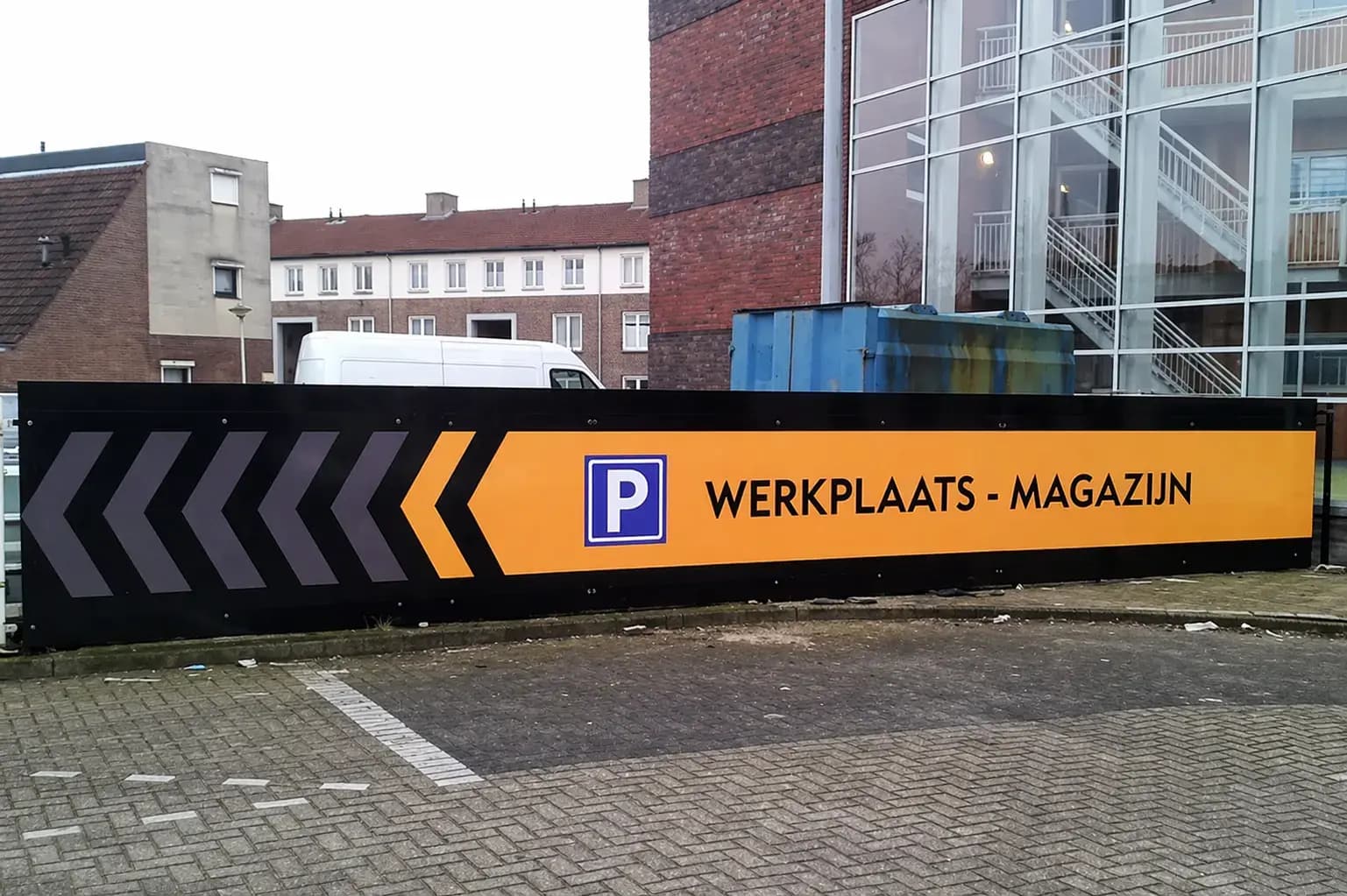 Groot zwart-geel wayfinding-bord met parkeerpictogram, linkerpijlen en de tekst WERKPLAATS - MAGAZIJN, geplaatst langs een parkeerterrein bij een gebouw.