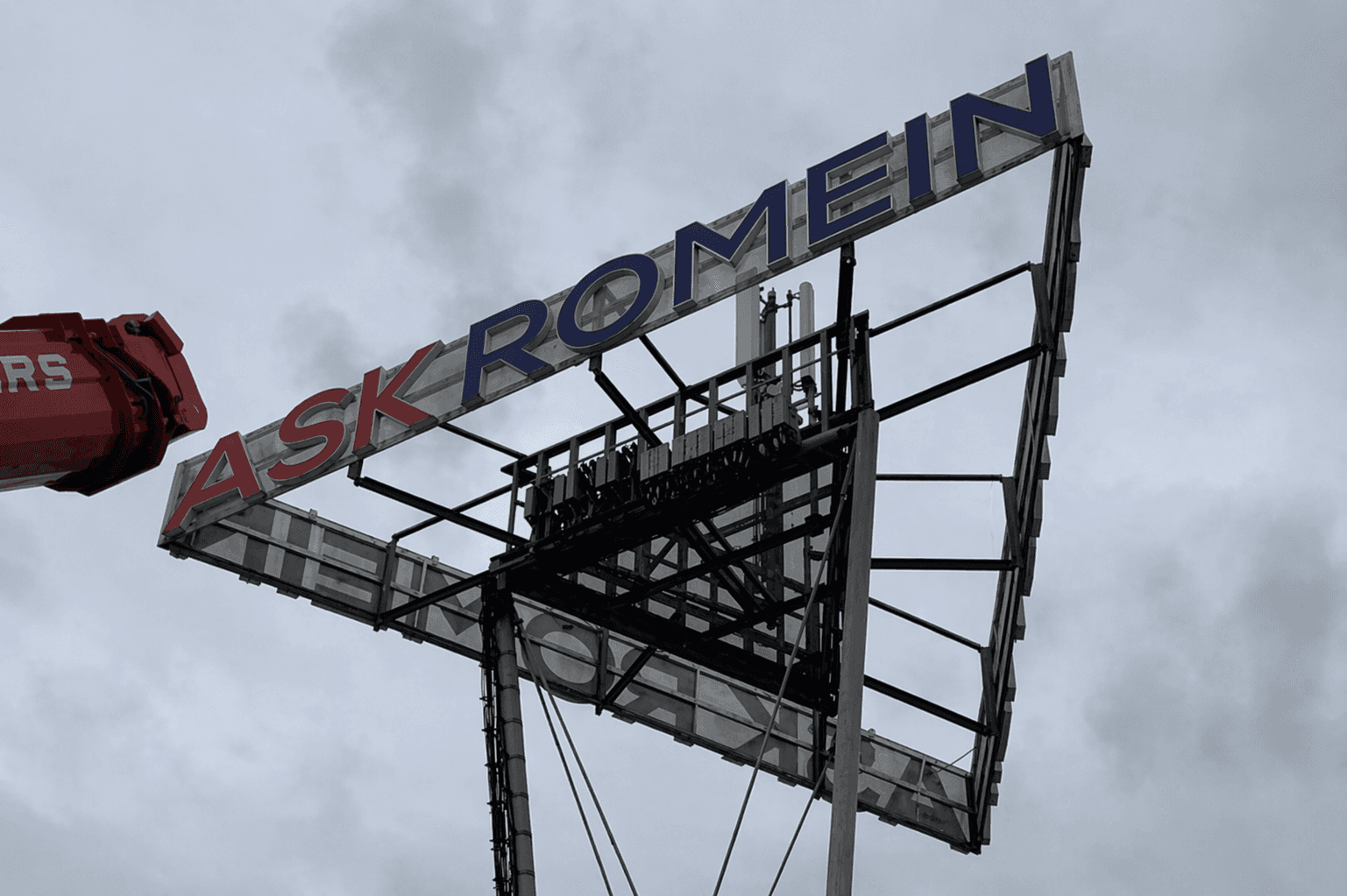 Grote hoog gemonteerde constructie met contourletters ASK ROMEIN in rood en blauw op een metalen frame, gefotografeerd tegen een bewolkte lucht.