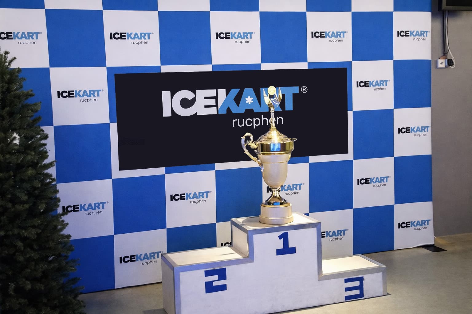 Blauw-witte fotowand met herhalend ICEKART Rucphen-logo, zwarte middenbanner en podium met beker voor een prijsuitreiking of evenement.