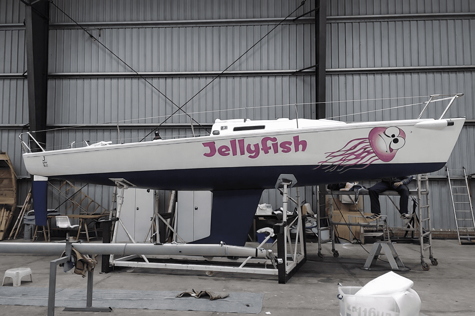 Witte zeilboot op bok in een werkplaats met roze bootbelettering ‘Jellyfish’ en een grote kwalillustratie op de romp.