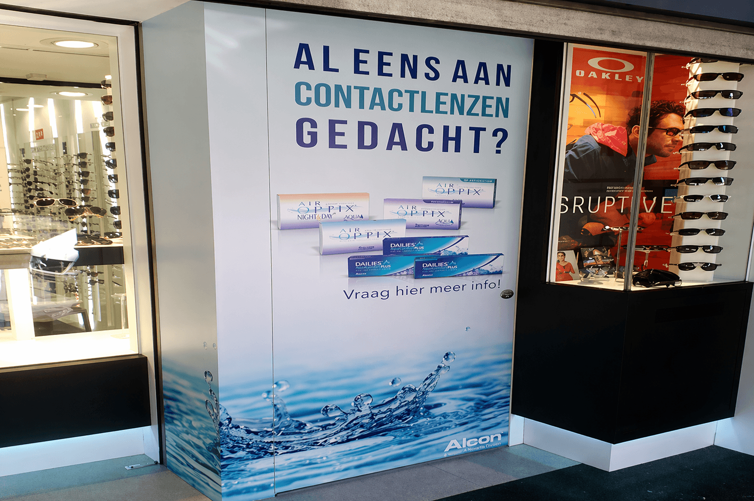 Grote full-colour wandreclame in een optiekzaak met contactlenzenpromotie, waterafbeelding en zichtbare productverpakkingen, naast een etalage met zonnebrillen en Oakley-display.