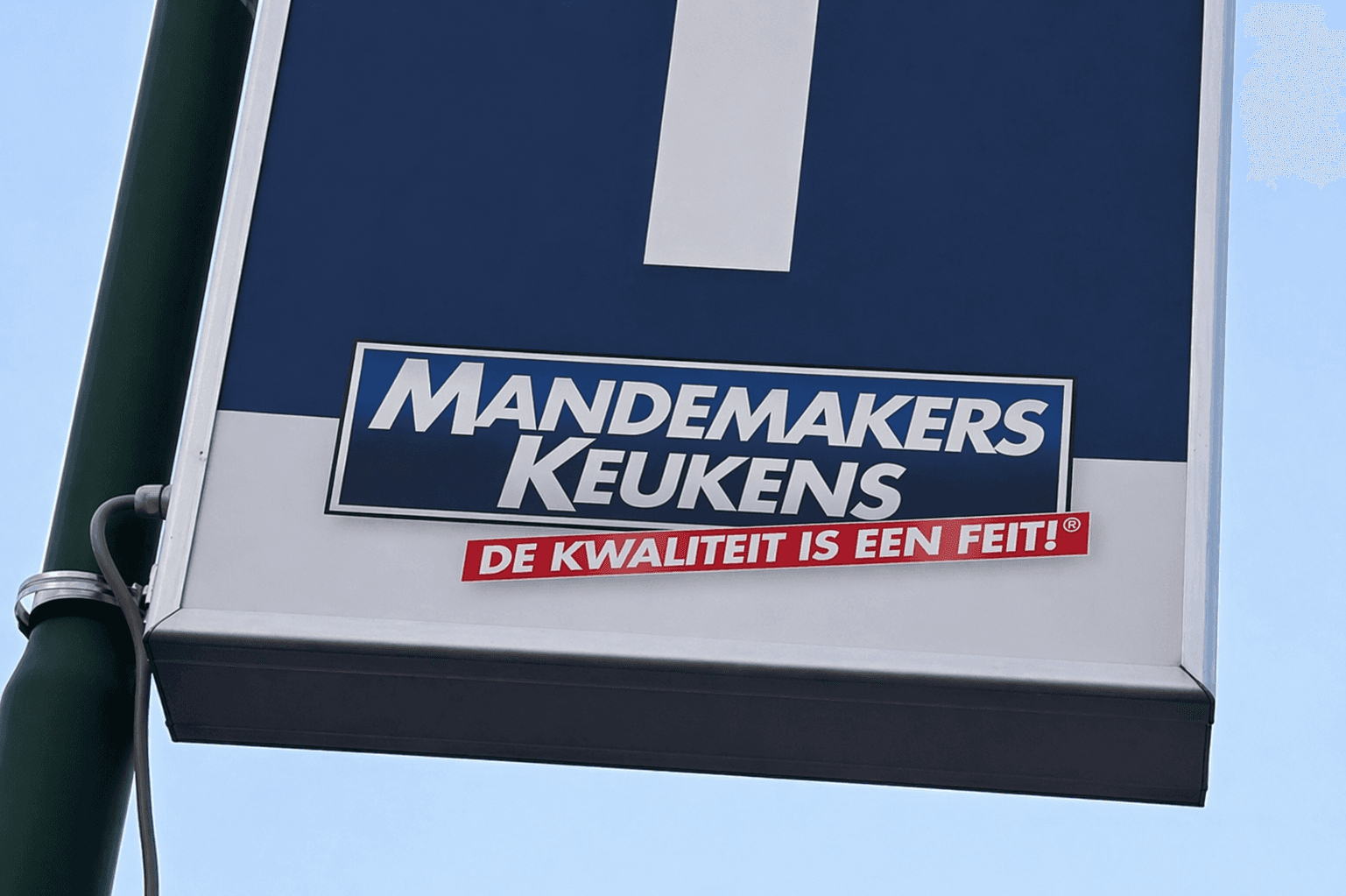 Blauw-wit wegwijzerbord op een groene paal met Mandemakers Keukens-logo en rode slogan, gefotografeerd tegen een heldere lucht.