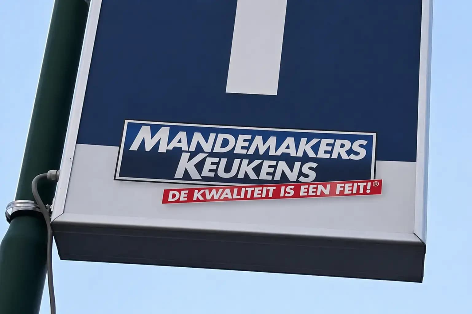 Blauw-wit wegwijzerbord op een groene paal met Mandemakers Keukens-logo en rode slogan, gefotografeerd tegen een heldere lucht.