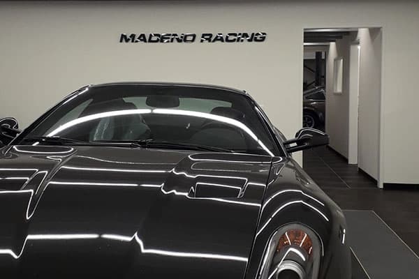 Interieur van een autoshop met zwarte sportwagen en zwarte wandbelettering "MADERO RACING" op een witte muur achter de auto.
