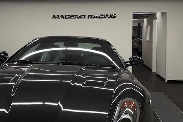 Interieur van een autoshop met zwarte sportwagen en zwarte wandbelettering "MADERO RACING" op een witte muur achter de auto.