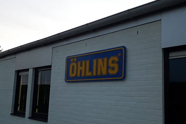 Witte bakstenen gevel met rechthoekig gevelbord van Öhlins in blauw en goud, gemonteerd als opvallende gevelreclame tussen zwarte kozijnen.
