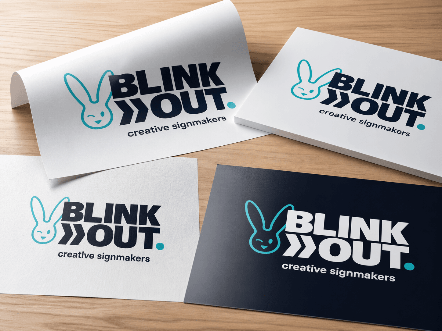 Logo-presentatie van BLINK OUT creative signmakers op verschillende drukwerktoepassingen, waaronder witte en donkerblauwe kaarten met contrasterende print op een houten ondergrond.