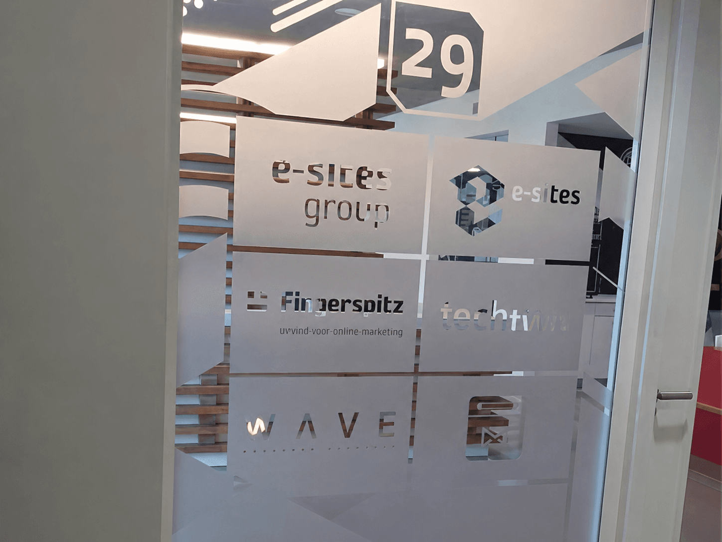 Glaswand met matte privacyfolie, uitgesneden logo’s en pijlvormen voor e-sites group, Fingerspitz en WAVE, toegepast als kantoorbelettering en wayfinding bij ruimte 29.