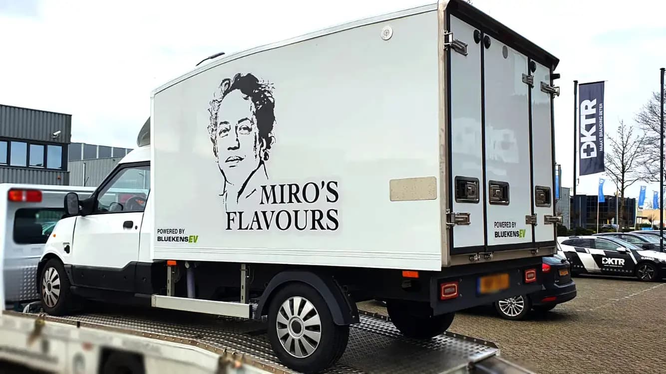 Witte bakwagen met zwarte voertuigbelettering voor Miro’s Flavours, voorzien van portretillustratie, merknaam op de laadbak en kleine sponsorvermelding op een bedrijventerrein.