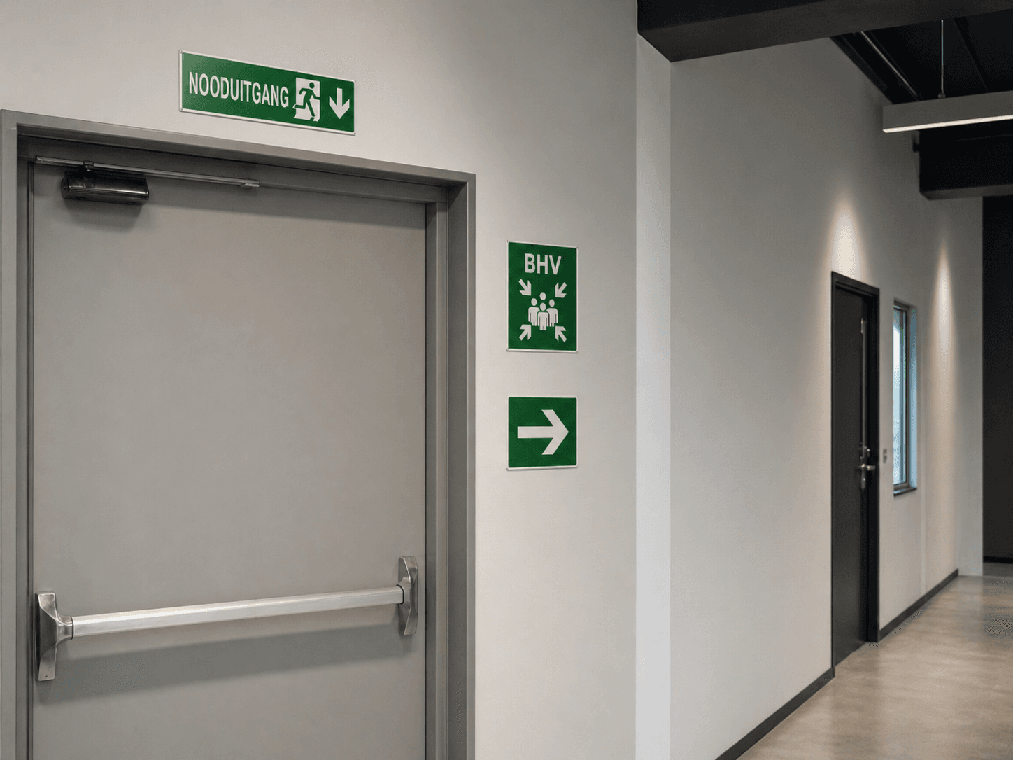 Groene nooduitgang- en BHV-veiligheidsborden met pijlmarkering bij een grijze vluchtdeur in een moderne gang.