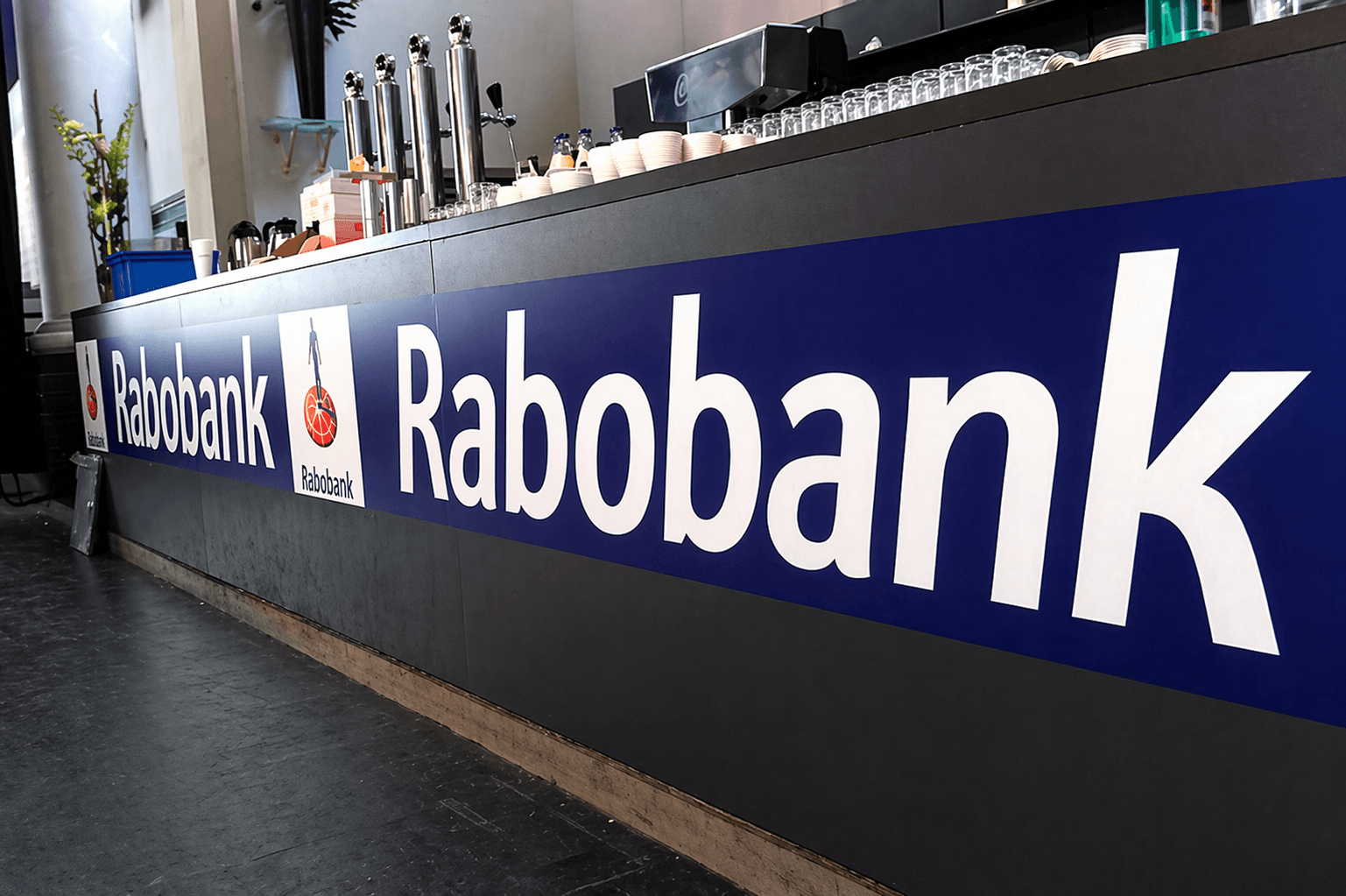 Blauwe Rabobank baliebelettering met witte logo’s en tekst op een donkere bar- of toonbankfront in een interieur.