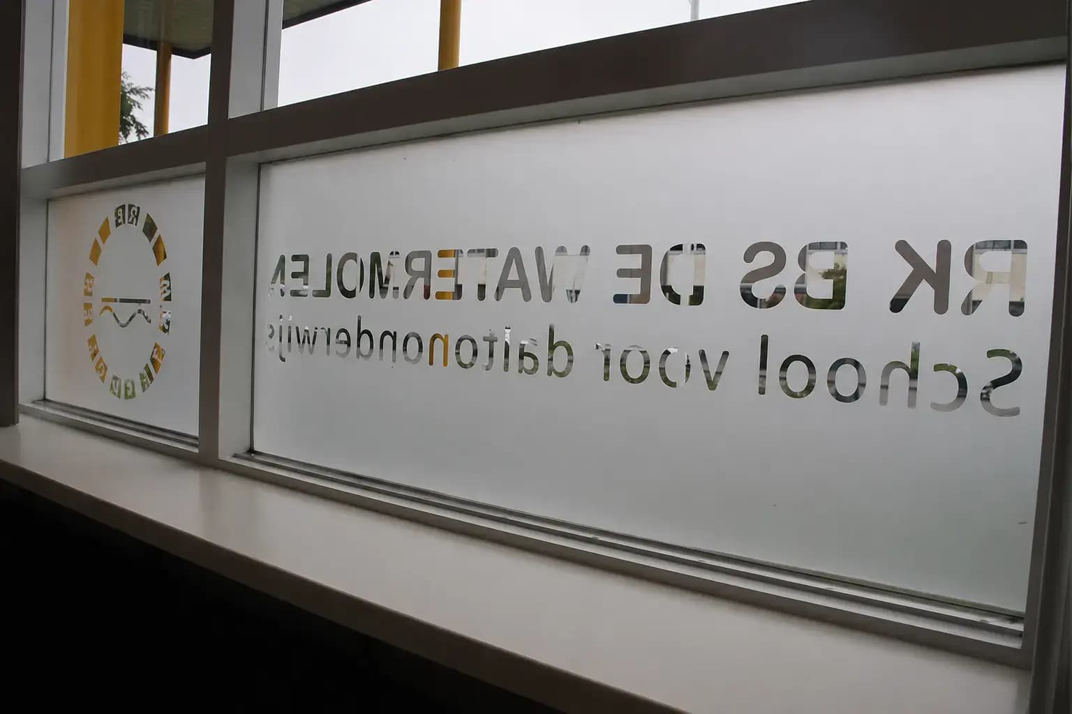 Glasfolie met uitgespaarde schoolnaam en rond logo op een raam, toegepast als raamdecoratie en privacyfolie.