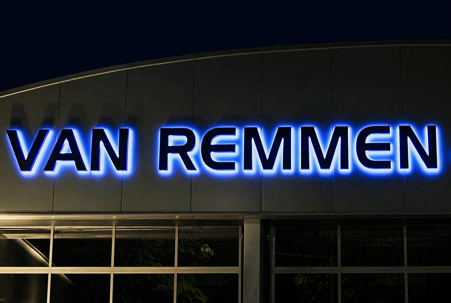 Blauwe halo-verlichte gevelletters met de tekst VAN REMMEN op een moderne bedrijfsgevel bij avond.
