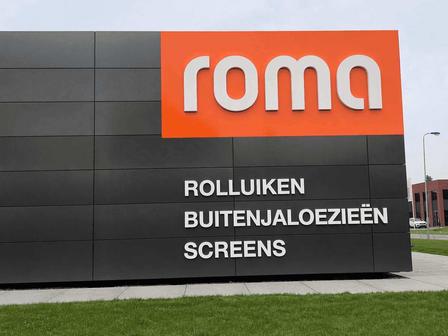 Moderne gevelreclame op zwarte gevelpanelen met oranje topvlak, witte 3D-letters van roma en productbelettering voor rolluiken, buitenjaloezieën en screens.