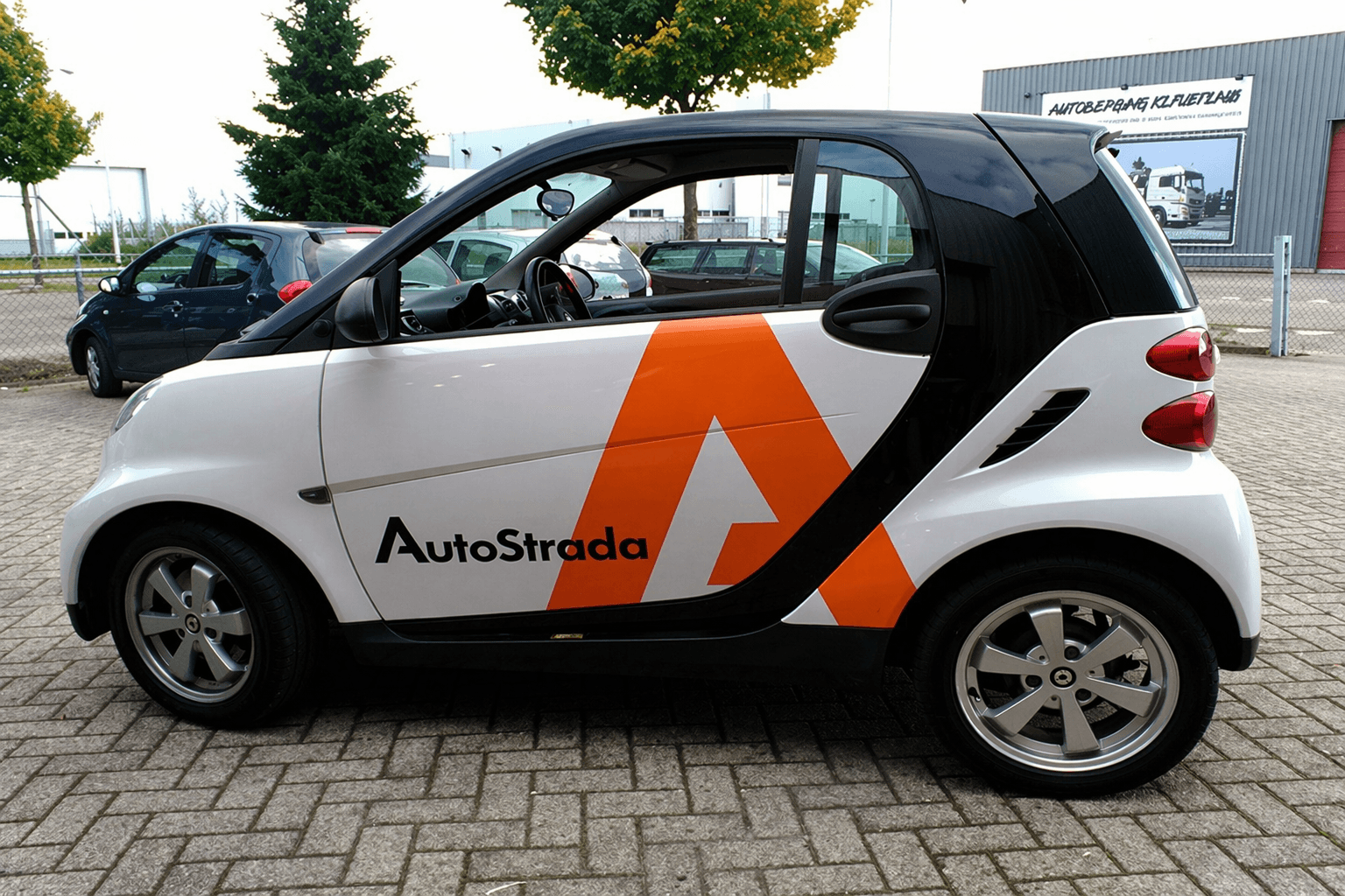 Zijaanzicht van een compacte witte stadsauto met zwarte panelen en opvallende oranje AutoStrada voertuigbelettering op de deur en achterzijde, geparkeerd op een bedrijventerrein.
