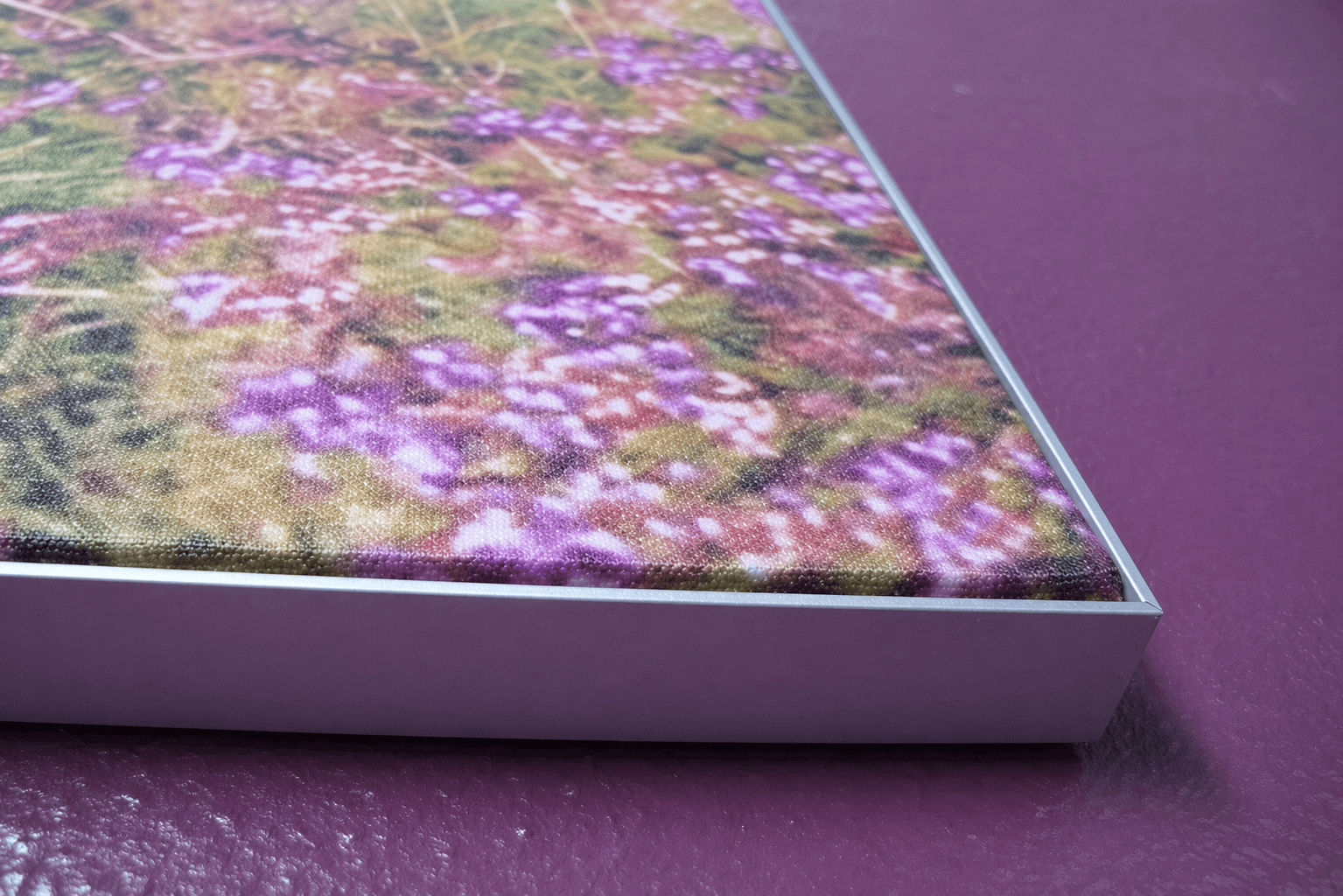 Close-up van een wanddecoratie met fotoprint van paarse bloemen in een strak aluminium frame, gemonteerd op een paarse structuurmuur.