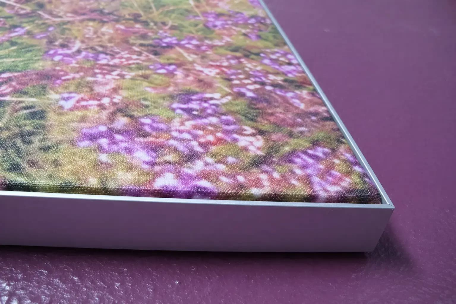 Close-up van een wanddecoratie met fotoprint van paarse bloemen in een strak aluminium frame, gemonteerd op een paarse structuurmuur.
