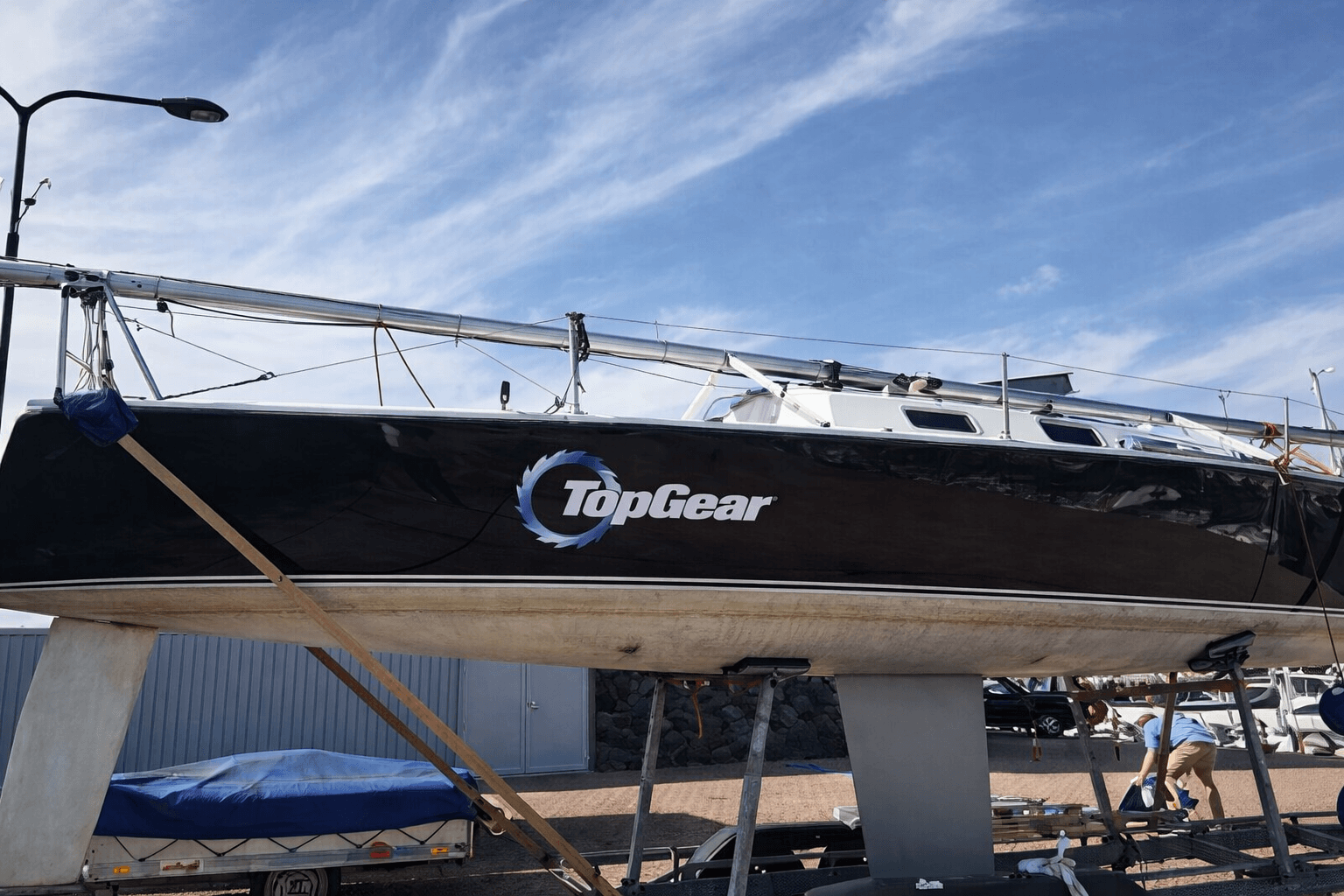 Zwarte zeilboot op bok met grote TopGear-belettering en ronde logosticker op de romp, gefotografeerd in een jachthaven onder een heldere lucht.