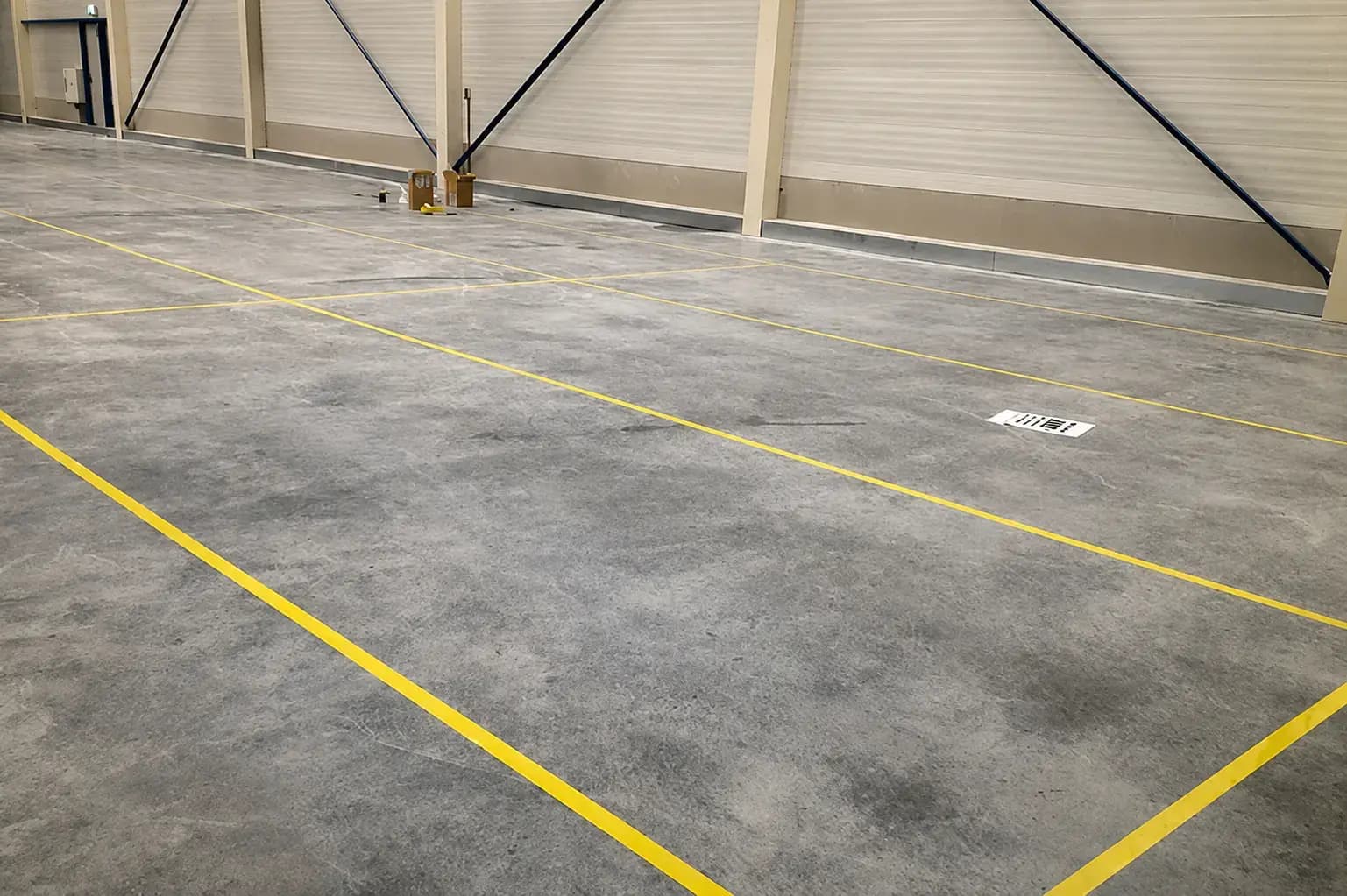 Gele vloermarkering op een betonnen bedrijfsvloer in een magazijnhal, met strakke belijning voor looproutes, werkzones en industriële wayfinding.