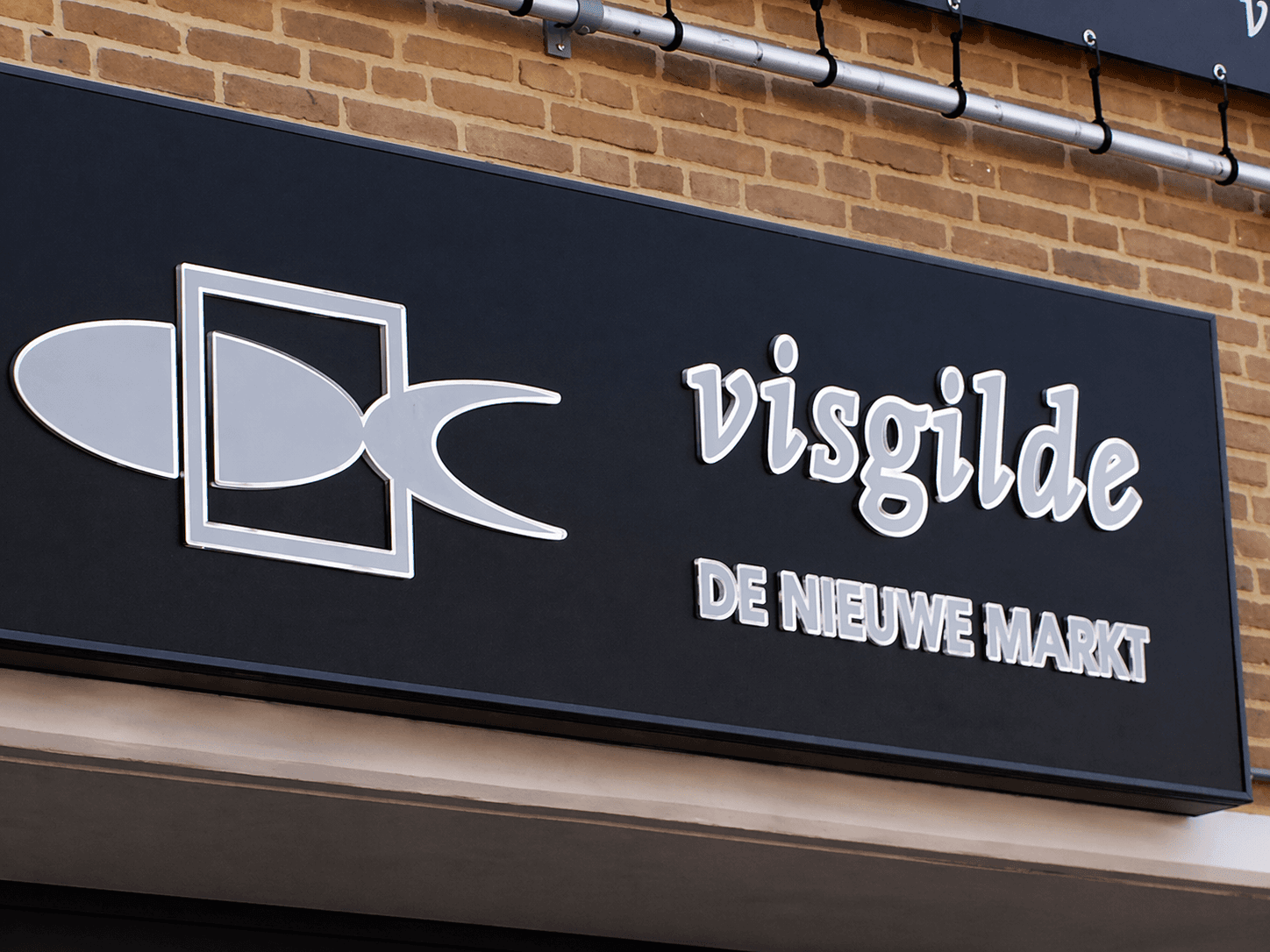 Zwart gevelbord met zilverkleurig logo en belettering van Visgilde De Nieuwe Markt, gemonteerd op een bakstenen winkelgevel.