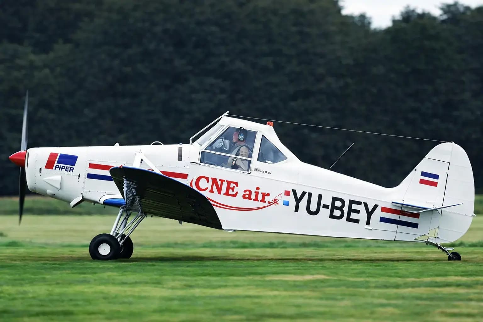Wit sportvliegtuig met Piper-logo, CNE air-belettering en grote registratiemarkering YU-BEY op de romp, gefotografeerd op een grasveld met bosrand op de achtergrond.