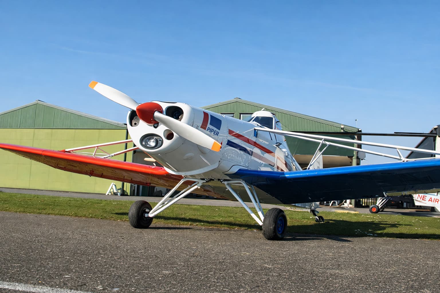Wit sportvliegtuig met Piper-logo, rood-blauwe striping en contrasterende vleugels, geparkeerd op een taxibaan voor hangars.