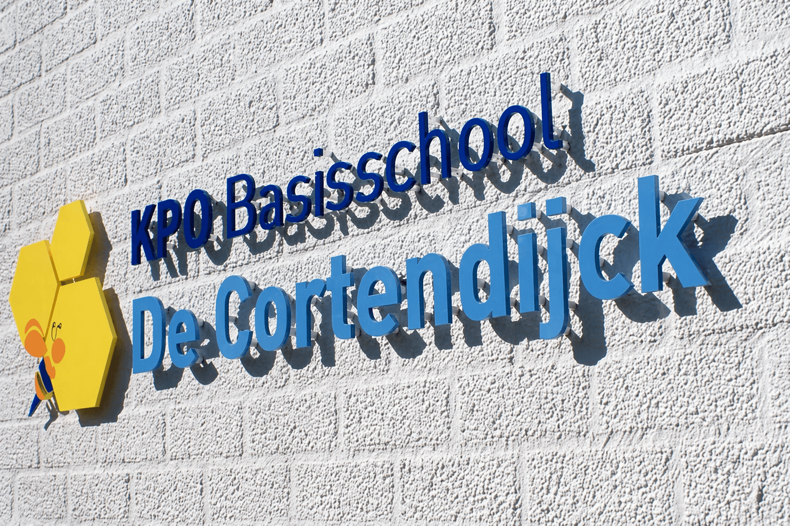 Witte gevel met blauwe 3D-belettering van KPO Basisschool De Cortendijck en een geel zeshoekig logo met bij, gemonteerd als opvallende schoolsigning.