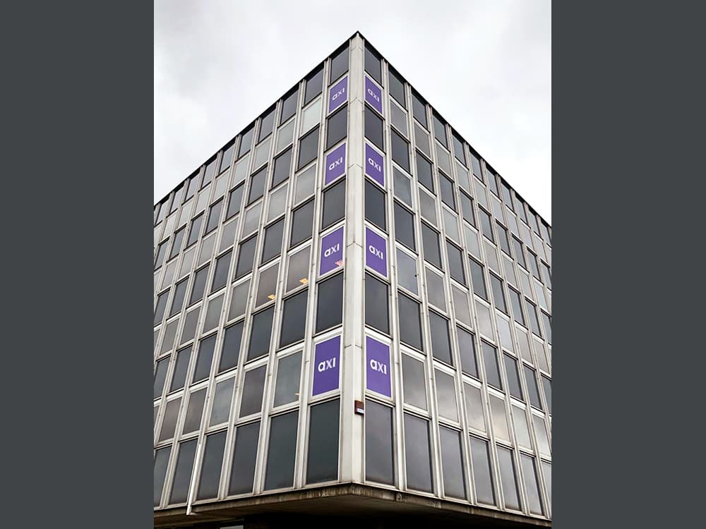 Hoekgevel van een modern kantoorgebouw met paarse raamstickers met axi-logo, verticaal aangebracht over meerdere verdiepingen als gevel- en raambelettering.