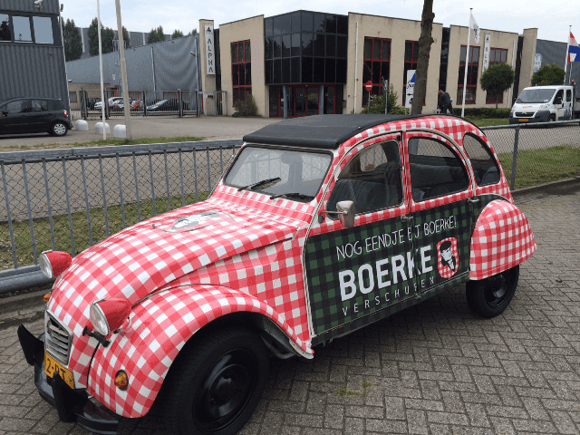 Klassieke auto met volledige voertuigwrap in rood-wit ruitpatroon en zwarte cabriokap, voorzien van groene zijbelettering met BOERKE-logo en promotionele tekst op een bedrijventerrein.