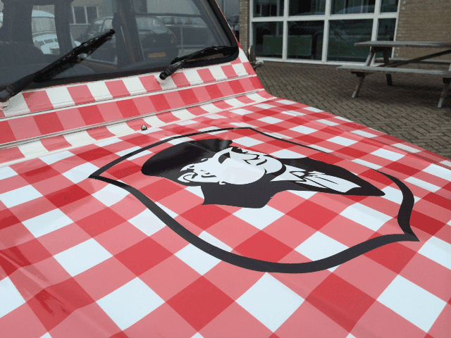 Close-up van de motorkap van een klassieke auto met rood-wit geruite voertuigwrap en groot zwart-wit illustratielogo op de voorzijde.