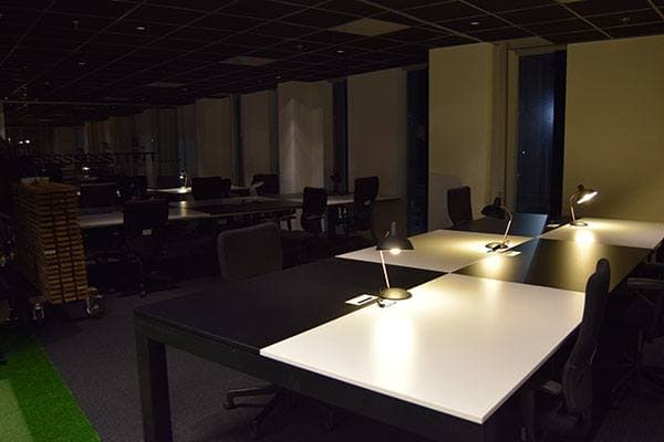 Donker kantoorinterieur met moderne werkplekken, bureaulampen op grote tafels en stoelen in een strak ingerichte open kantoorruimte.