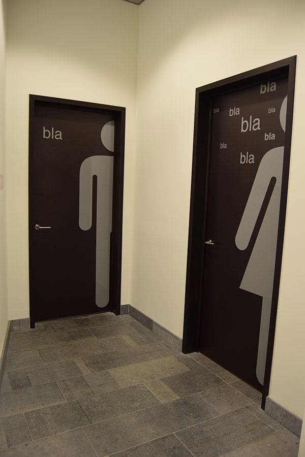 Twee donkere binnendeuren met grote grijze deurfolie in figuurvorm en typografische belettering, geplaatst als decoratieve wayfinding in een modern kantoorinterieur.
