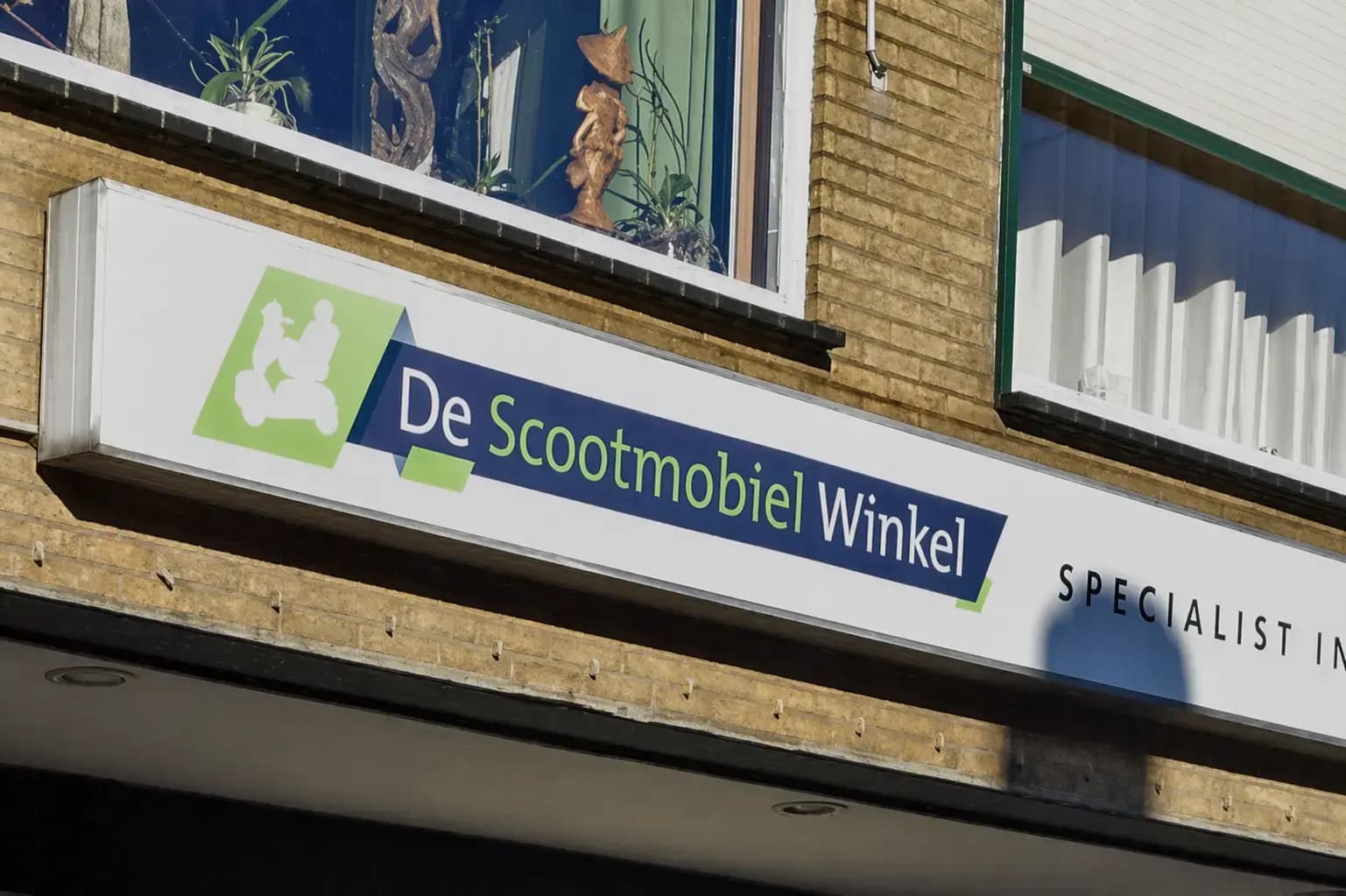 Wit gevelbord met groen-blauw logo en belettering van De Scootmobiel Winkel, gemonteerd op een bakstenen winkelgevel onder de ramen.