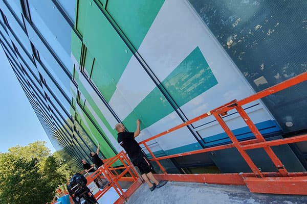 Montage van full-colour raamfolie op een glazen gevel met grote grafische vlakken, uitgevoerd vanaf oranje hoogwerkers aan een modern kantoorgebouw.
