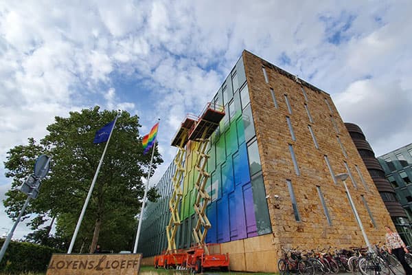 Modern kantoorgebouw met glazen hoekgevel voorzien van regenboogkleurige raamfolie, gefotografeerd tijdens montage met hoogwerkers en vlaggen voor het pand.