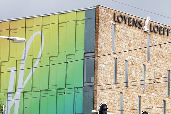 Hoekgevel van een modern kantoorgebouw met zwarte gevelletters "Loyens & Loeff" op de natuurstenen gevel en groen-gele raamfolie met grote witte vorm op de glazen zijgevel.