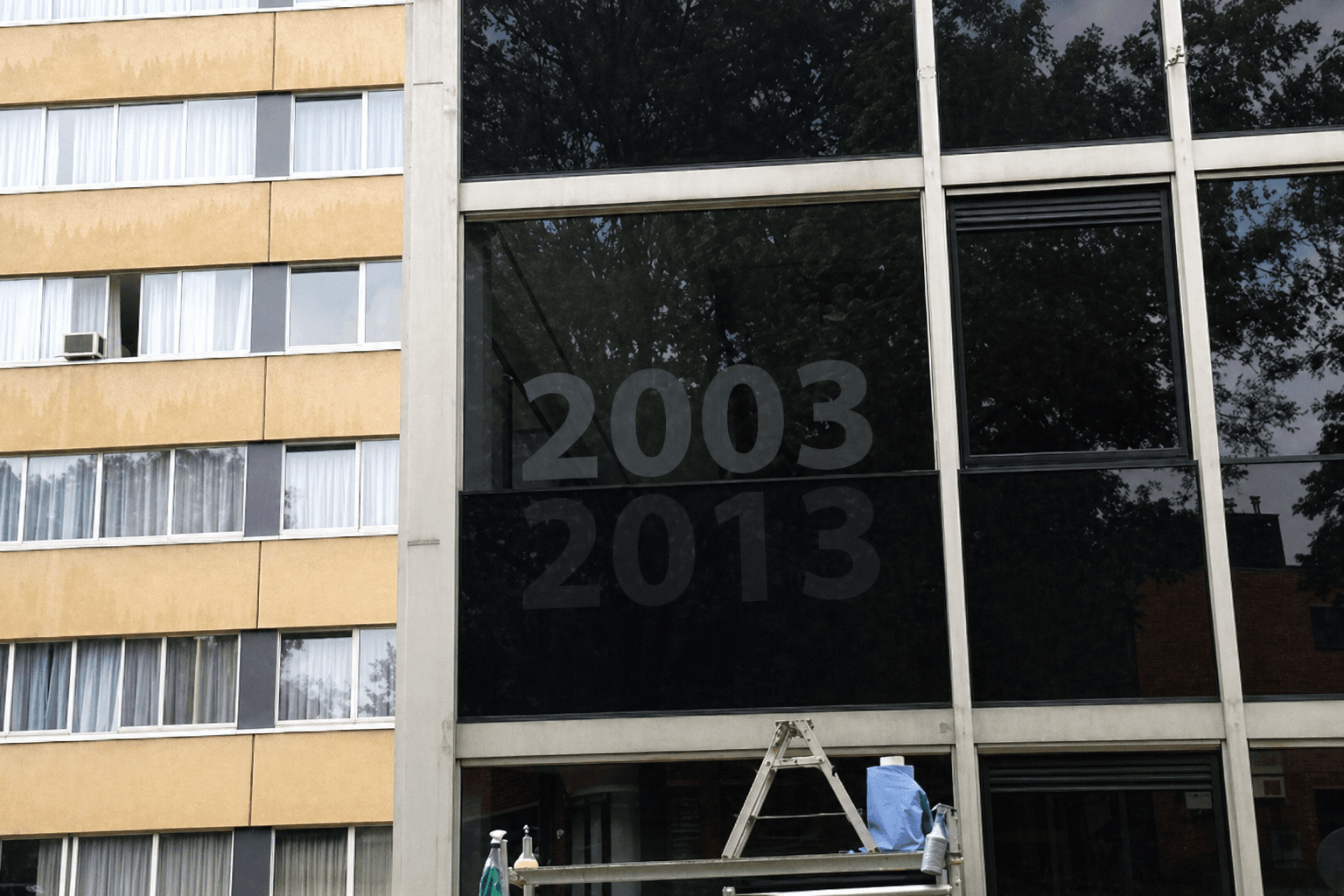 Donkere raamfolie met uitgespaarde jaartallen 2003 en 2013 op een glazen gevel, aangebracht boven een werkplatform met ladder.
