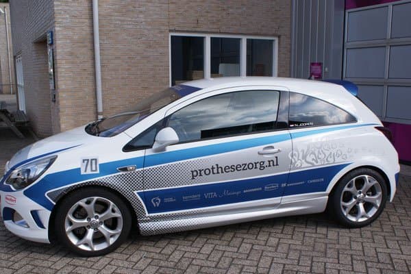 Zijaanzicht van een witte personenauto met blauwe en grijze voertuigbelettering, prothesezorg.nl op het portier en sponsorlogo’s langs de onderzijde, geparkeerd op een bedrijventerrein.