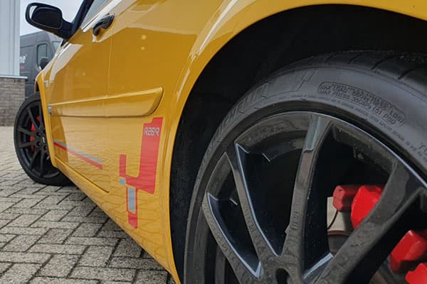 Close-up van een gele personenauto met sportieve voertuigbelettering op de dorpel, zwarte lichtmetalen velg en rode remklauw.