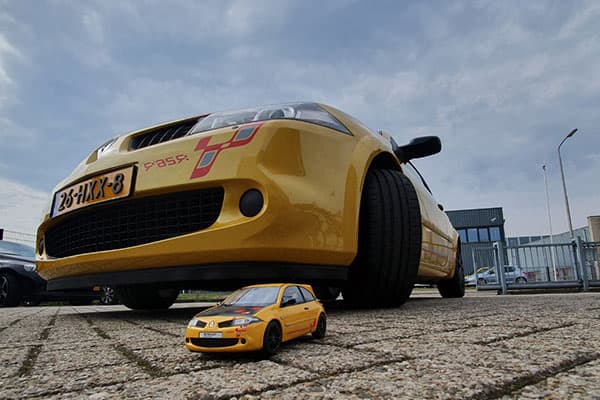 Gele personenauto met sportieve voertuigbelettering en rode striping op de voorzijde, gefotografeerd vanaf laag standpunt met een bijpassend miniatuurmodel op de bestrating.