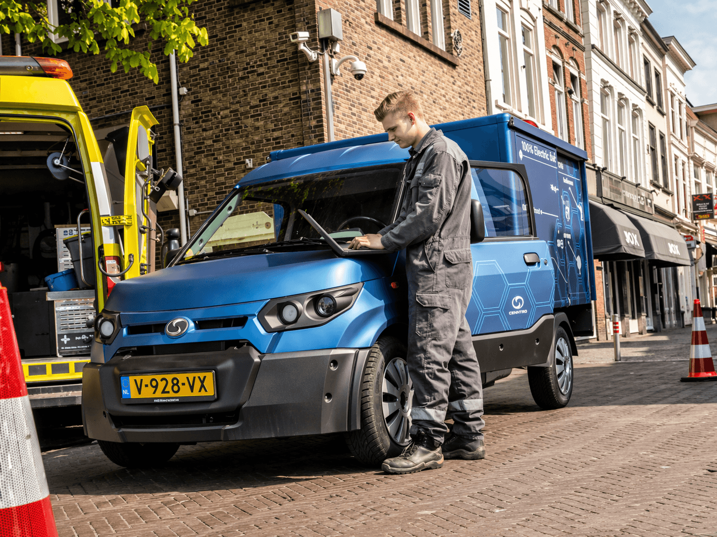 Blauwe bedrijfswagen met full-colour voertuigbelettering en bestickering geparkeerd in een winkelstraat, terwijl een monteur met laptop naast het voertuig staat.